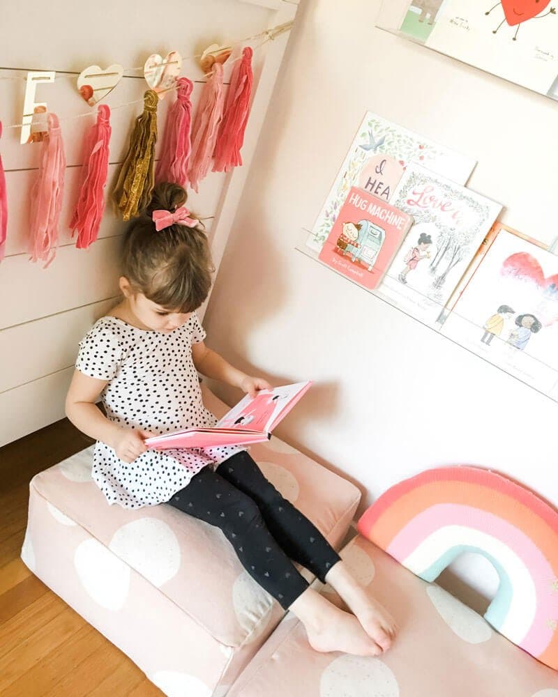 A girl sitting on a pink pillow reading a Valentine's Day picture book.