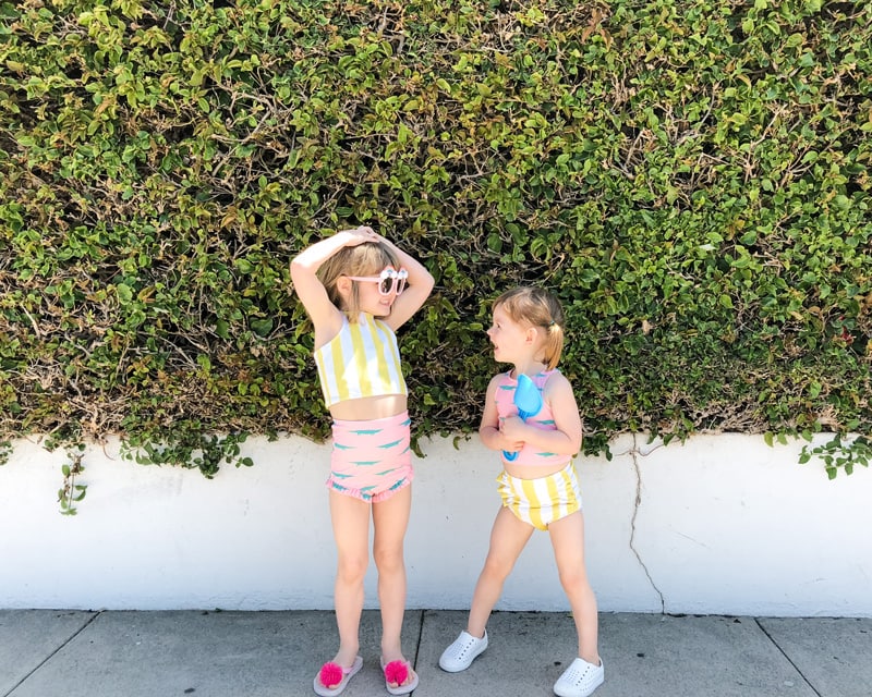 two smiling little girls wearing swimsuits and standing on a sidewalk