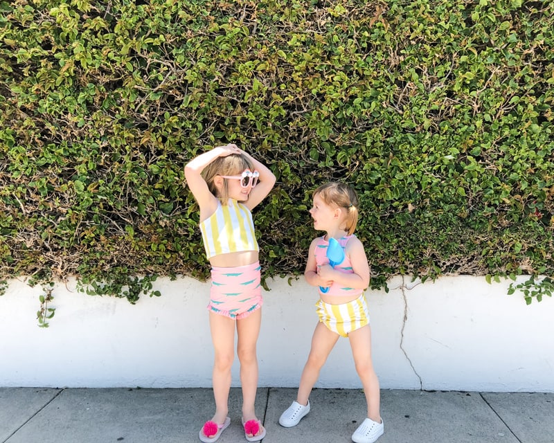 two smiling little girls wearing swimsuits and standing on a sidewalk 