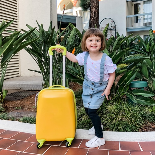 A young girl standing with yellow suitcase in front of a hotel.