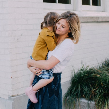 A mother holding her toddler standing in front of a white building
