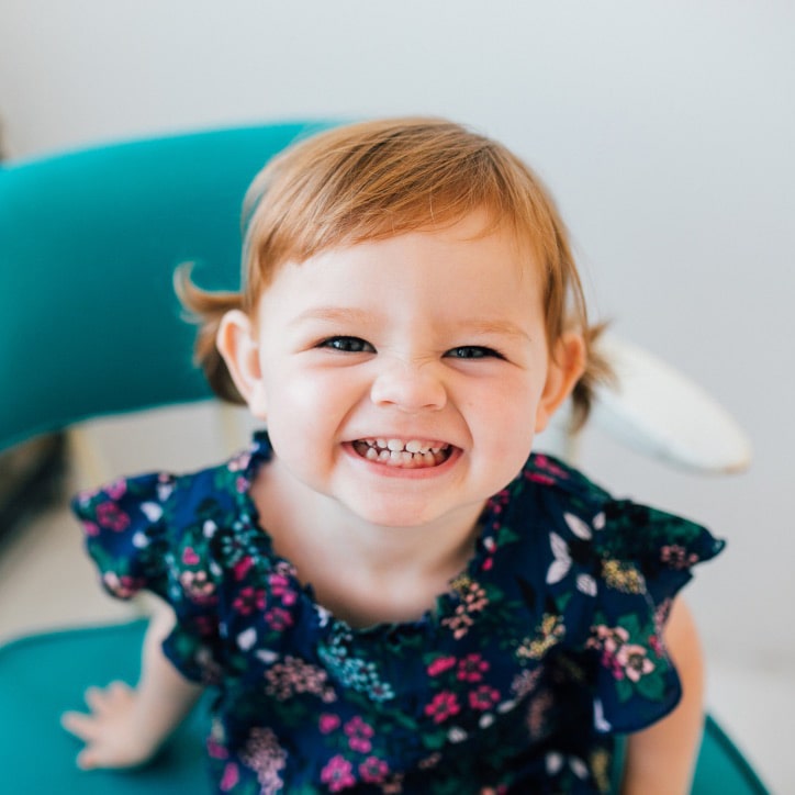 A red haired toddler with a floral dress sitting in a turquoise chair