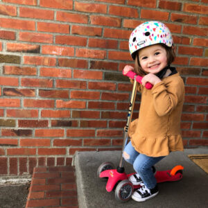 A young girl riding a scooter with a helmet and smiling