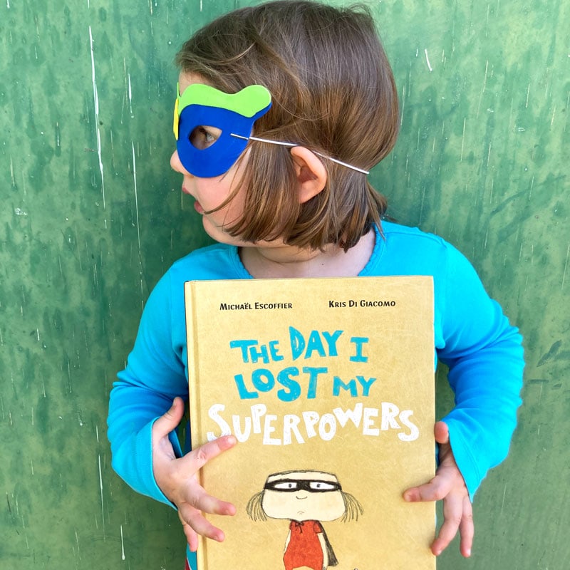 girl with short brown hair in a blue shirt and superhero eye mask holding the book titled "The Day I Lost My Superpowers"